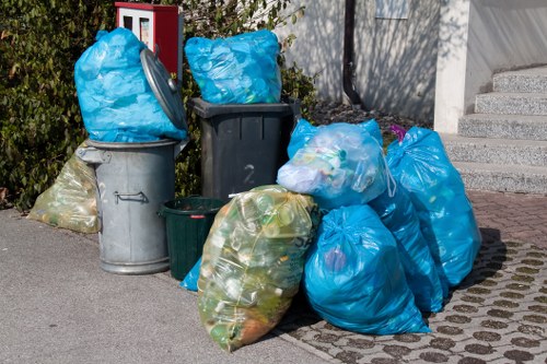 A collection of various waste collection bins and bags positioned on a paved outdoor surface, including a large metallic grey bin, a black plastic wheelie bin, and several blue and green plastic rubbish bags filled with assorted household waste. The bags are tied at the top, some leaning against the bins, while others are placed directly on the ground. The scene is set adjacent to a building with steps leading up to an entrance, and a garden area with greenery and purple flowers is visible in the background. The environment appears to be part of a residential or commercial parking area, with natural daylight illuminating the scene, indicating daytime in Vauxhall. This image reflects typical rubbish collection practices and would be relevant in the context of waste or rubbish removal services such as those offered by Garden Clearance Vauxhall.