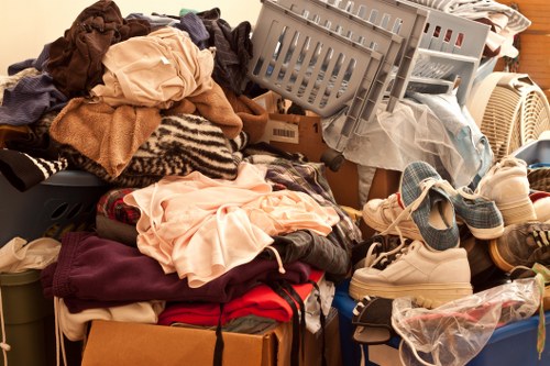 A disorganized pile of various used clothing items, including shirts, trousers, and jackets, is spread across a surface with some pieces spilling over onto the floor. Among the clothing, a grey plastic crate filled with more fabric and miscellaneous refuse is partially visible, leaning to one side. A pair of sneakers and other footwear are stacked or placed haphazardly near the pile, with some shoes wrapped in plastic. The background includes a beige or light-colored wall and a portable heater or similar appliance among the clutter. The scene appears to be an indoor setting, possibly a storage area or a room awaiting clearance, illustrating a typical example of rubbish or unwanted items that Garden Clearance Vauxhall might help remove from residential or commercial properties in Vauxhall or nearby areas in London.