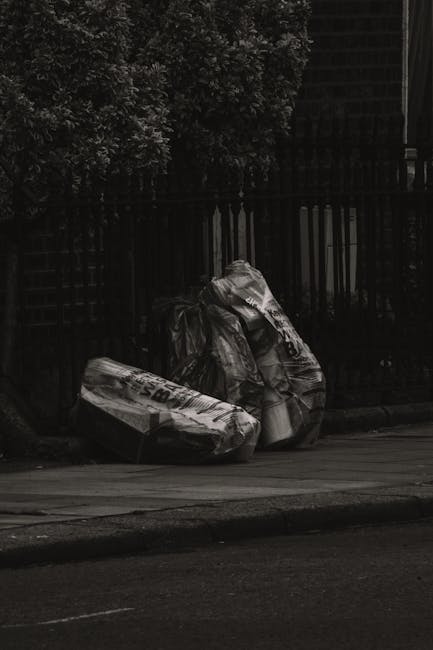 A low-angle photograph of a pile of mixed rubbish discarded on a street surface near a stone wall. The debris includes black plastic trash bags, a yellow plastic container, and a weathered, light brow
